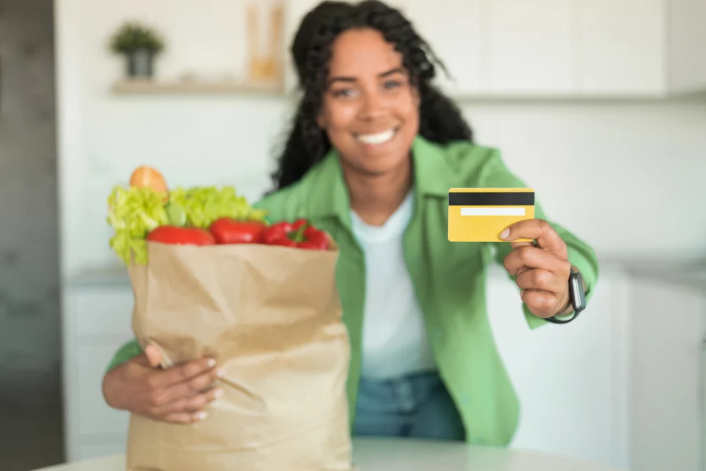 Colaboradora sorridente segura cartão de benefícios corporativos do Novo PAT 2026 ao lado de sacola de papel com alimentos frescos na cozinha.